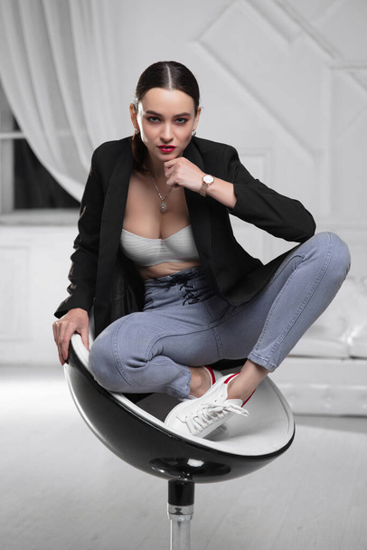 Beautiful lady wearing a jacket, bra and jeans sitting on an armchair - Fotó, kép
