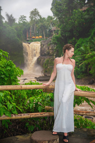 Beautiful woman posing against the background of muddy waters of Tegenungan waterfall during the rainy season, Bali island, Indonesia - 写真・画像