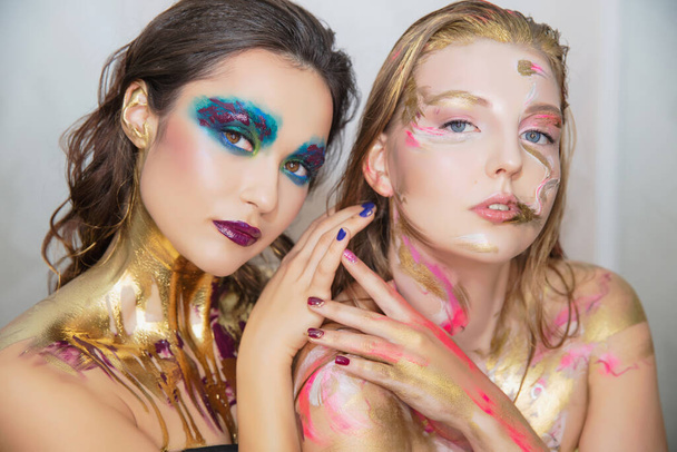 Twee schattige jonge vrouwen met creatieve make-up op het gezicht poseren in de studio. - Foto, afbeelding