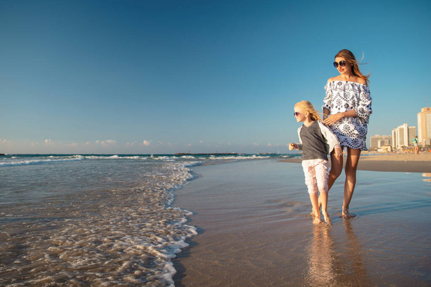Bonita mamá con una hija caminando descalza a lo largo del oleaje en la playa de arena - Foto, Imagen