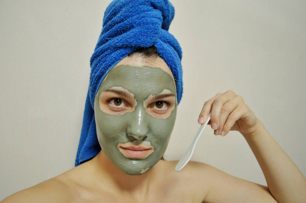 へらを持つ女性は彼女の頭に青いタオルで彼女の顔に粘土マスクを適用します - 写真・画像