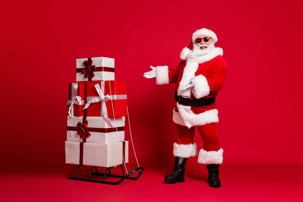 Full length photo of retired old man white beard recommending sled many giftbox best choice wear santa x-mas costume coat sunglass headwear black boots isolated red color background - 写真・画像