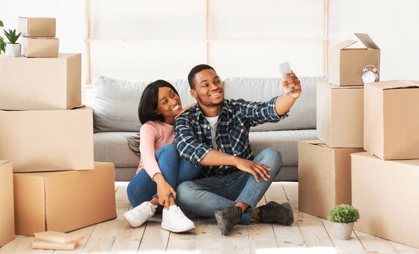 Sonriente chico negro y su novia mudándose a su propia casa juntos, tomando selfie entre cajas de cartón, panorama - Foto, Imagen