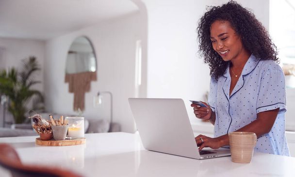 Woman Wearing Pyjamas In Kitchen Making Online Credit Card On Laptop - Φωτογραφία, εικόνα