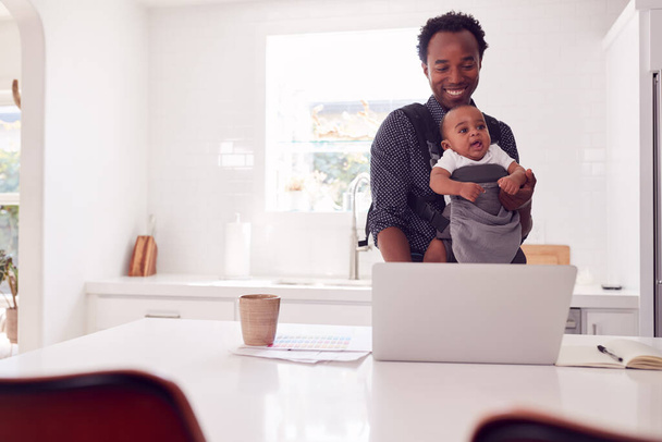 Father With Baby Daughter In Sling Multi-tasking Working From Home On Laptop - 写真・画像