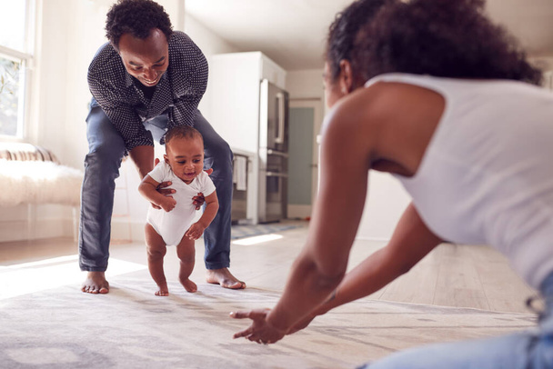 Parents Encouraging Smiling Baby Daughter To Take First Steps And Walk At Home - Fotó, kép