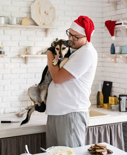 Kerst koken. Jongeman in kerstmuts houdt zijn hond in zijn armen in de keuken - Foto, afbeelding