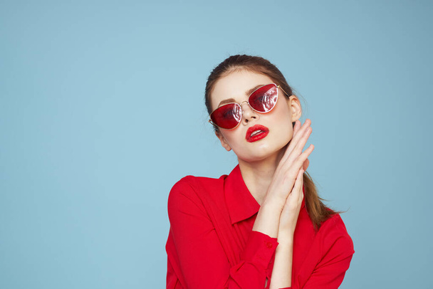 Woman in red shirt on blue background holds hands near face and sunglasses red lips makeup model - Foto, Imagem