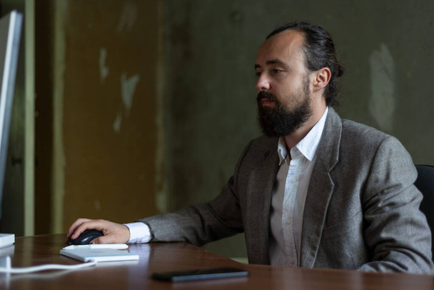 Ofisteki bilgisayar masasında takım elbiseli sakallı genç bir iş adamı oturuyor. Bir bilgisayar faresi, grafiksel tablete bir liste yazar. Ofis yenileme neşeli işadamı ofiste dizüstü bilgisayarla oturuyor. - Fotoğraf, Görsel
