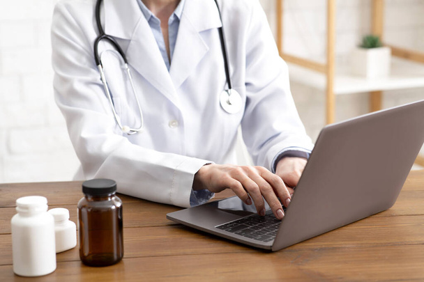 Таблетки, лечение и защита здоровья. Взрослая женщина-врач в белом халате со стетоскопом, работающая на ноутбуке - Фото, изображение