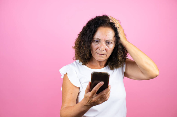 Mulher de meia idade vestindo camisa branca casual em pé sobre fundo rosa isolado conversando, surpreso e olhando para o telefone - Foto, Imagem