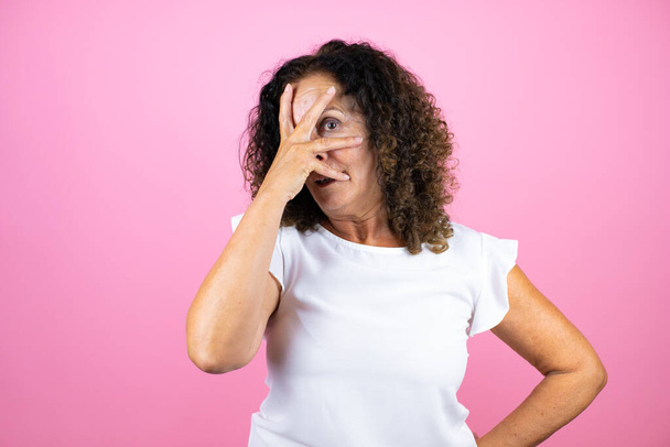 Keski-ikäinen nainen päällään rento valkoinen paita seisoo yli eristetty vaaleanpunainen tausta kurkistaa shokki kattaa kasvot ja silmät käsin, katselee sormien kautta hämmentynyt ilme - Valokuva, kuva