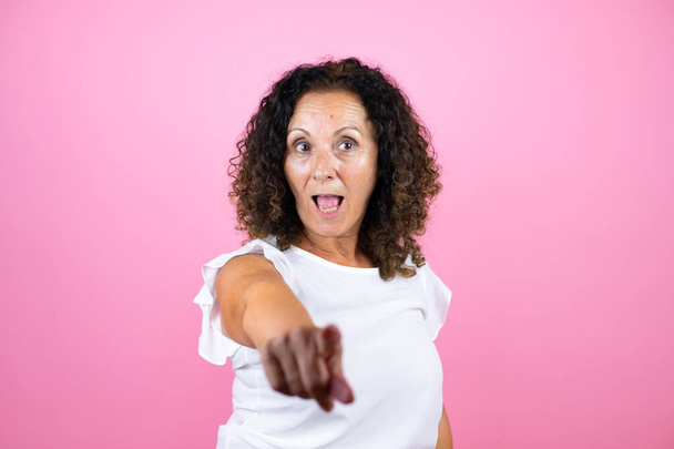 Žena středního věku v neformální bílé košili stojící nad izolovaným růžovým pozadím ukazující prstem překvapeně dopředu, s otevřenými ústy užaslý výraz, něco na přední straně - Fotografie, Obrázek