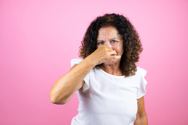Женщина средних лет в обычной белой рубашке, стоящей на изолированном розовом фоне, пахнет чем-то вонючим и отвратительным, невыносимым запахом, затаив дыхание пальцами на носу - Фото, изображение