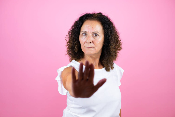 Middelbare leeftijd vrouw het dragen van casual wit shirt staan over geïsoleerde roze achtergrond serieus en doen stoppen met zingen met palm van de hand. - Foto, afbeelding