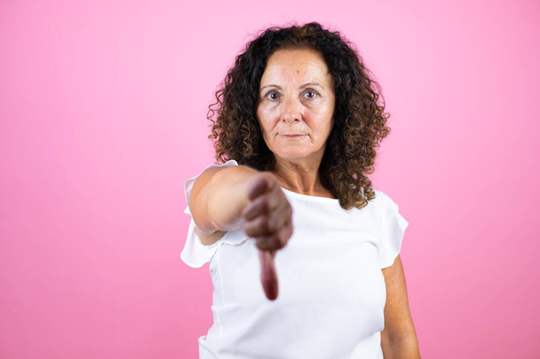 Mujer de mediana edad con camisa blanca casual de pie sobre un fondo rosa aislado con la cara enojada, signo negativo que muestra aversión con el pulgar hacia abajo - Foto, Imagen