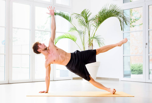 Hombre haciendo un ejercicio de yoga de tablón lateral para fortalecer sus músculos centrales, brazo y muñeca en la vista lateral en un gimnasio de alta clave en un concepto de salud y fitness - Foto, imagen