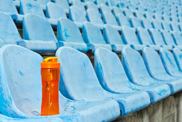 Na stadionu je spousta volných míst. Oranžová láhev s vodou na sedadle. Pij pro sportovce. Plastová sedadla jsou uspořádána v řadě. Špinavá a poškrábaná modrá sedadla ventilátoru. - Fotografie, Obrázek