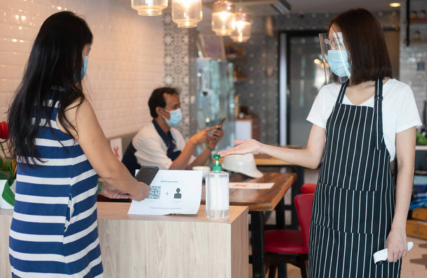 Asyalı garson kadın yüz maskesi takıyor ve elinde kızılötesi termometre tutuyor ve müşterilere restorana girmeden önce el alkolü dezenfektanı kullanmalarını öneriyor (kahve dükkanı) ). - Fotoğraf, Görsel