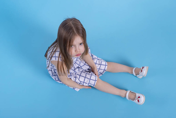 Volledig portret van een beetje gefrustreerd of beledigd meisje met pruillip zittend op blauwe achtergrond in studio en kijkend naar camera, bovenaanzicht - Foto, afbeelding
