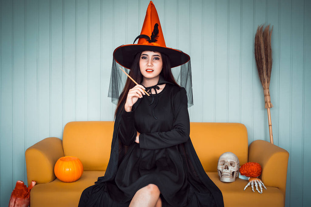 Attraktive schöne asiatische Frau als Hexe verkleidet, Zauberstab in der Hand und auf gelbem Sofa sitzend, Halloween-Feiertage im Filmton.   - Foto, Bild