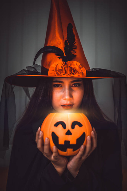 Attractive beautiful Asian woman dressed as a witch holding jack-o-lantern on hand, Halloween holidays in cinematic dark tone.   - Photo, Image