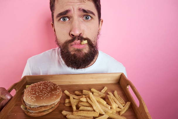 Man with a tray of food in his hand hamburger fries and fast food calorie pink background portrait close-up - Photo, Image