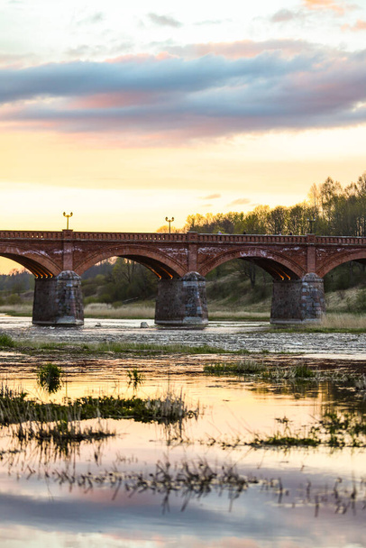 ベンタ川の上の古い歴史的なレンガ造りの橋の深夜の夕日の景色。ラトビアで撮影. - 写真・画像