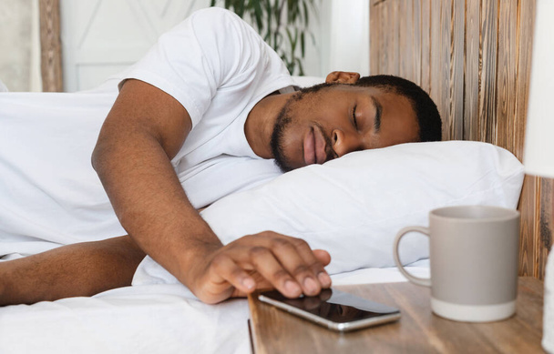 ベッドに寝そべっているスマートフォンの目覚まし時計を通して眠るアフリカ人男性 - 写真・画像