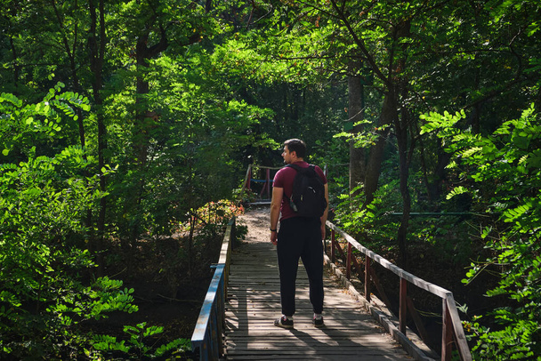 Un hombre con una mochila en un puente de madera. Hombre excursionista de pie en un puente entre el verde parque de verano. Caminar en el bosque verde de verano - Foto, imagen