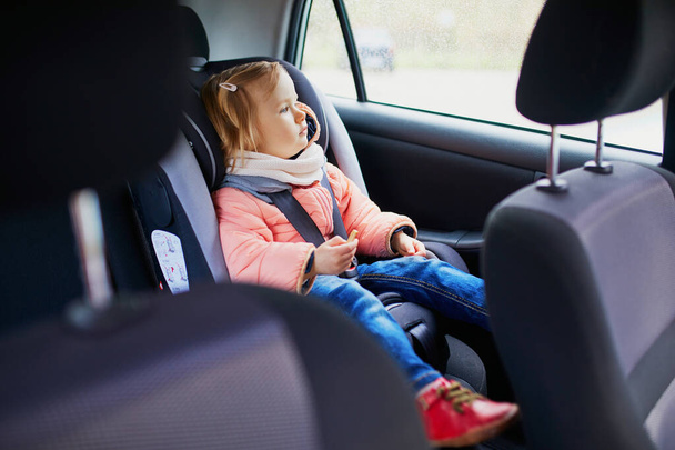 Adorable toddler girl in modern car seat eating cookie. Little kid traveling by car. Child safety on the road. Trip with a baby - Photo, image