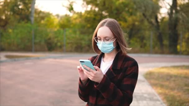 Nainen lääketieteellisessä suojapeitteessä ja lasit käyttää älypuhelinta ulkona. - Materiaali, video