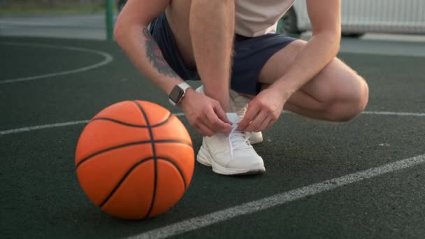 Tipo irreconocible atando cordones en zapatillas de deporte en cuclillas junto a la pelota de baloncesto - Metraje, vídeo