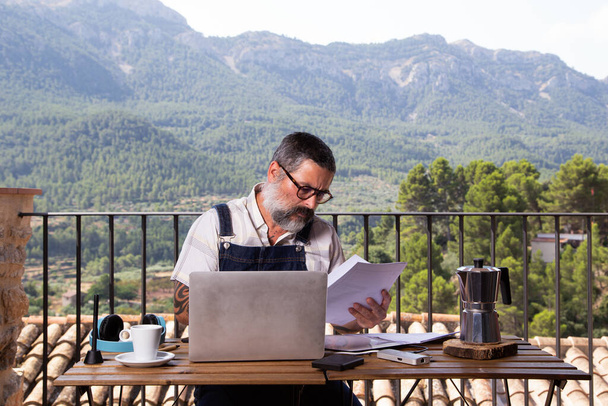 Man met een witte baard zittend met een laptop op het terras in een groot raam. Man met behulp van laptop, winkelen of online werken van thuis uit buiten. Onafhankelijk en zelfstandig - Foto, afbeelding