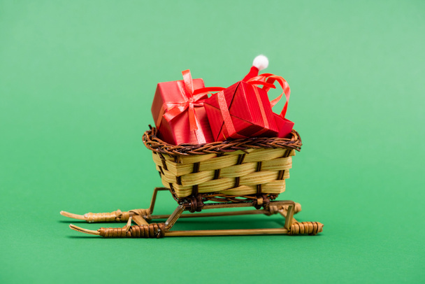 Kırmızı hediye kutuları ve hasır sepette Noel Baba şapkası yeşil arka planda dekoratif kızak - Fotoğraf, Görsel