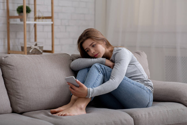 Porträt einer depressiven jungen Frau, die mit dem Smartphone in der Hand auf dem Sofa sitzt - Foto, Bild