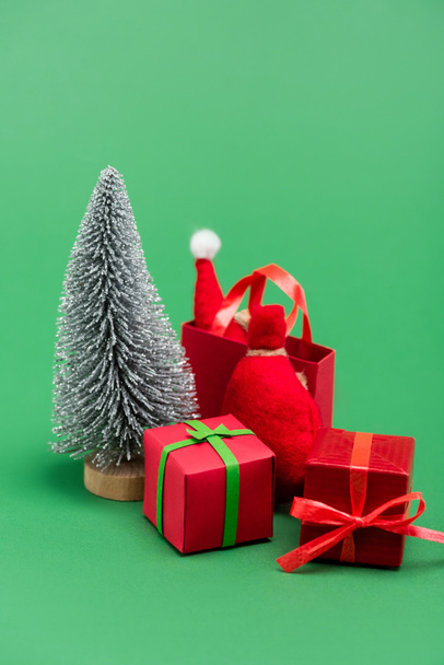 dekorativní stříbrný vánoční stromek v blízkosti dárkových krabic, pytel a nákupní taška se Santou klobouk na zelené - Fotografie, Obrázek