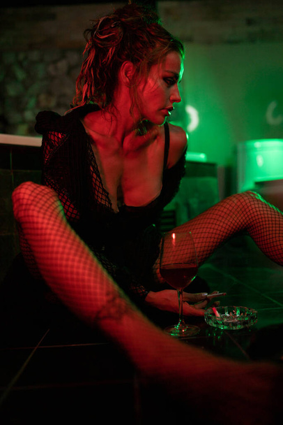 Sexy Frau sitzt auf dem Boden im Badezimmer, raucht und trinkt Wein. Rotes und grünes Licht. Filmdreh - Foto, Bild