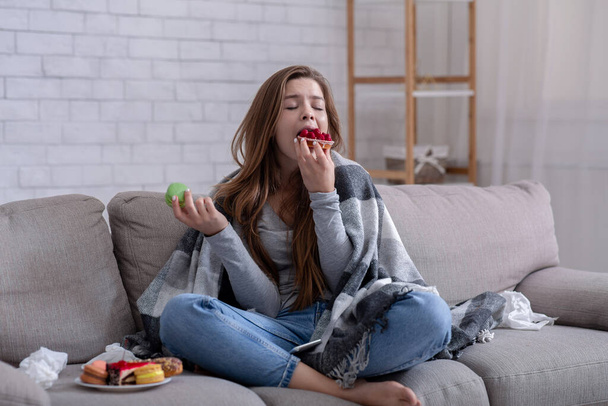 Sad young lady coping with depression or stress by eating sweets on sofa at home - Photo, image