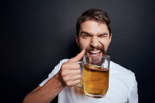 man with a mug of beer in a white t-shirt emotions lifestyle drunk on a dark isolated background - Photo, image