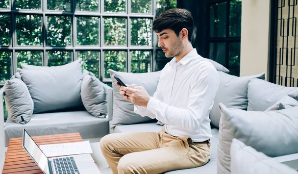 Side view of pensive ethnic male entrepreneur chatting on mobile phone and sitting at table on sofa with laptop while working remotely in cafe - 写真・画像