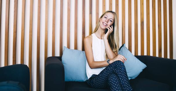 Young stylish blond haired woman looking confidently at camera while resting on blue couch in lounge and chatting via mobile phone - 写真・画像