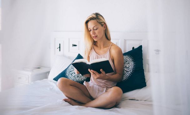 Serious woman dressed in casual wear interested on plot of bestseller relaxing in home interior during free time, young Caucasian female reader making research of useful information in literature book - Фото, изображение