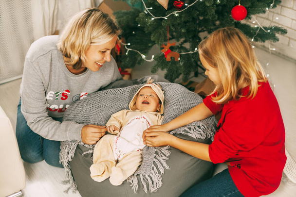 Un bébé joyeux se trouve à côté du sapin de Noël et sourit à sa mère et sa sœur. Une mère et une fille jouent avec un bébé dans une chambre décorée pour Noël. Vacances, Nouvel an, Noël - Photo, image