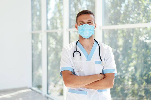 Confident male doctor wearing medical mask with arms crossed in hospital background - Photo, image