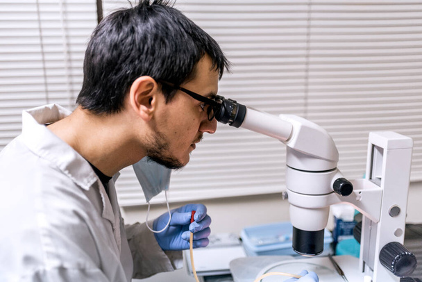Фотография мужчины-ученого в маске для лица с помощью микроскопа в его лаборатории. - Фото, изображение