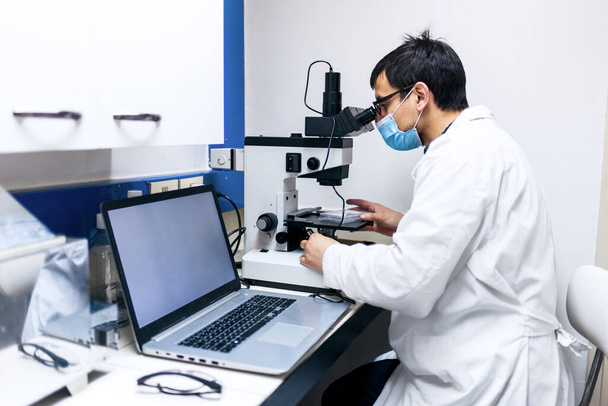 Foto stock di scienziato di sesso maschile con maschera facciale utilizzando il computer e una microscopia nel suo laboratorio. - Foto, immagini
