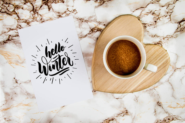Caffè su un supporto a forma di cuore su uno sfondo di marmo con pezzo di carta bianco e decorazioni per le vacanze invernali con testo Ciao inverno  - Foto, immagini
