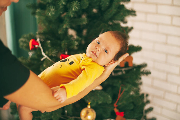 Крупним планом дитина в руках матері дивиться на камеру на тлі прикрашеної ялинки. Подія, Різдво та Новий рік. Простір для тексту
 - Фото, зображення