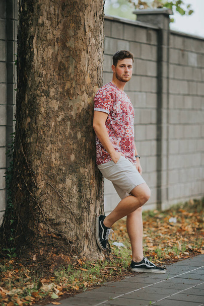 A young Caucasian male in a red patterned shirt and shorts leaning against the tree - Photo, Image